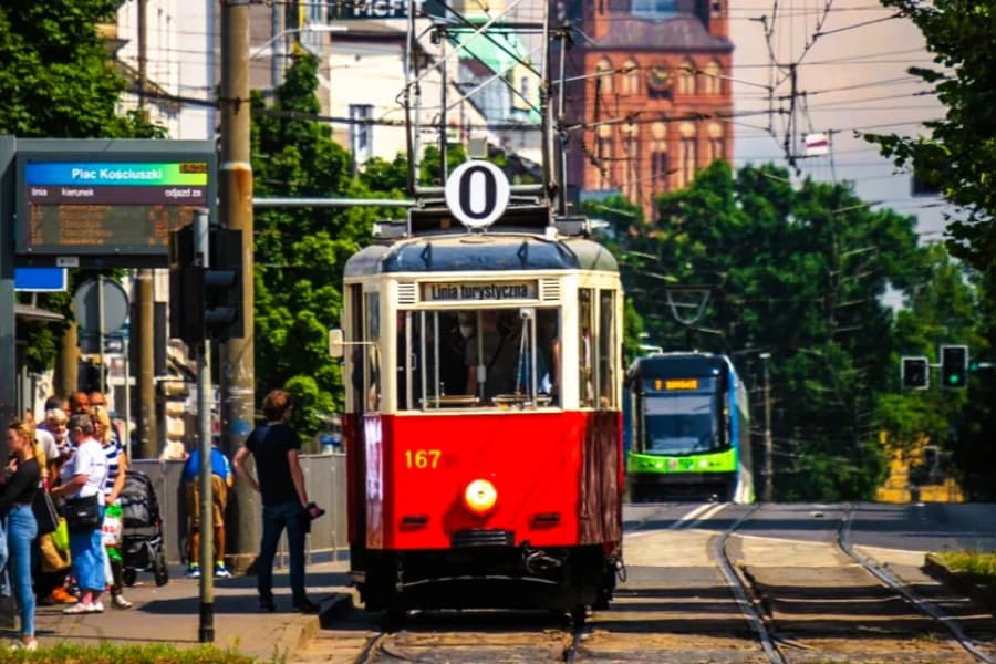 Przedostatni wakacyjny weekend z turystyczną linią 0 Czerwony tramwaj turystyczny linii 0. Z lewej strony przystanek z grupą ludzi.
